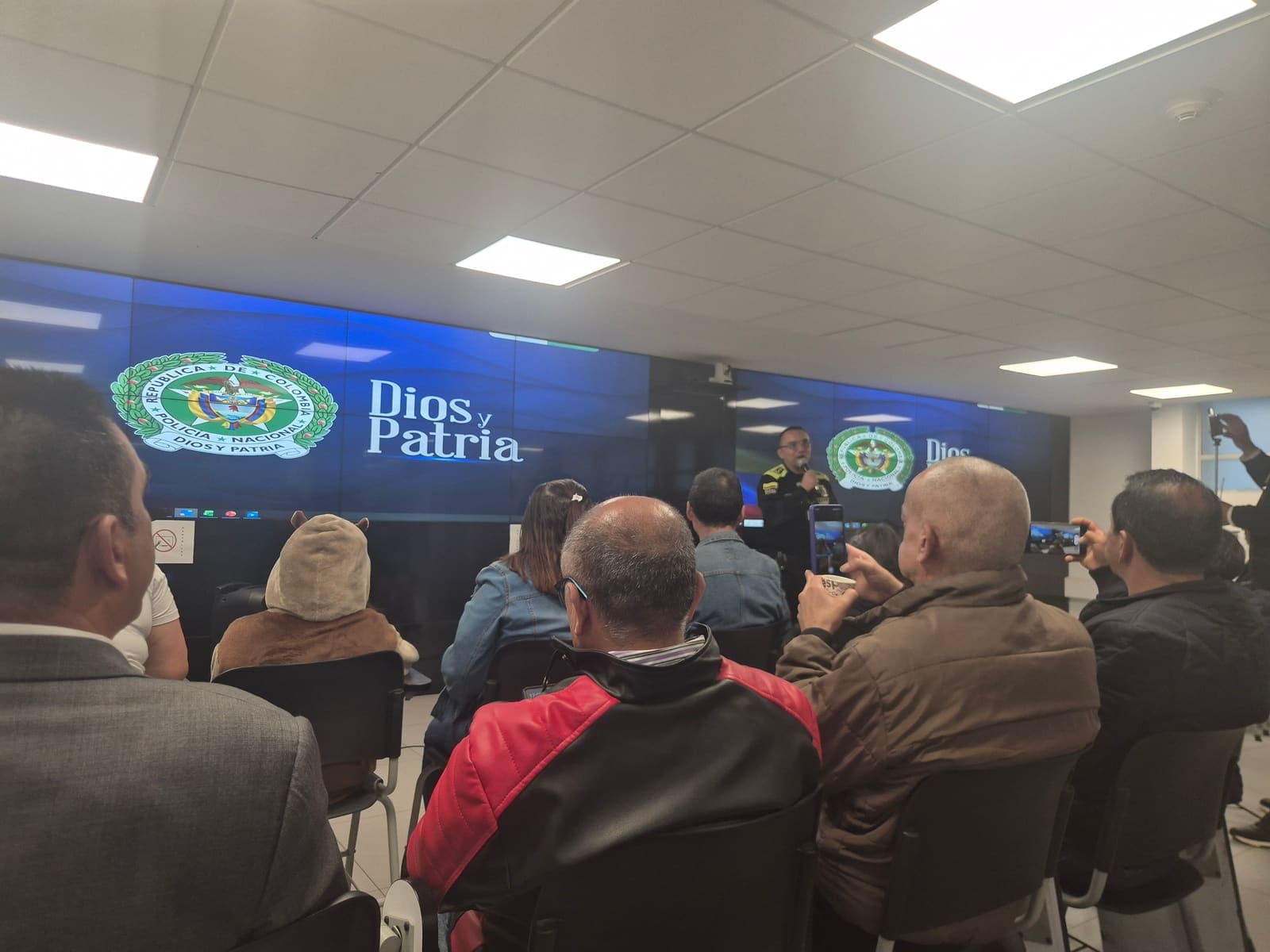 Coordinadores de las Zonas Seguras de Kennedy reunidos en la Estación de Policía, escuchando la intervención de un oficial frente a la pantalla institucional con el lema Dios y Patria
